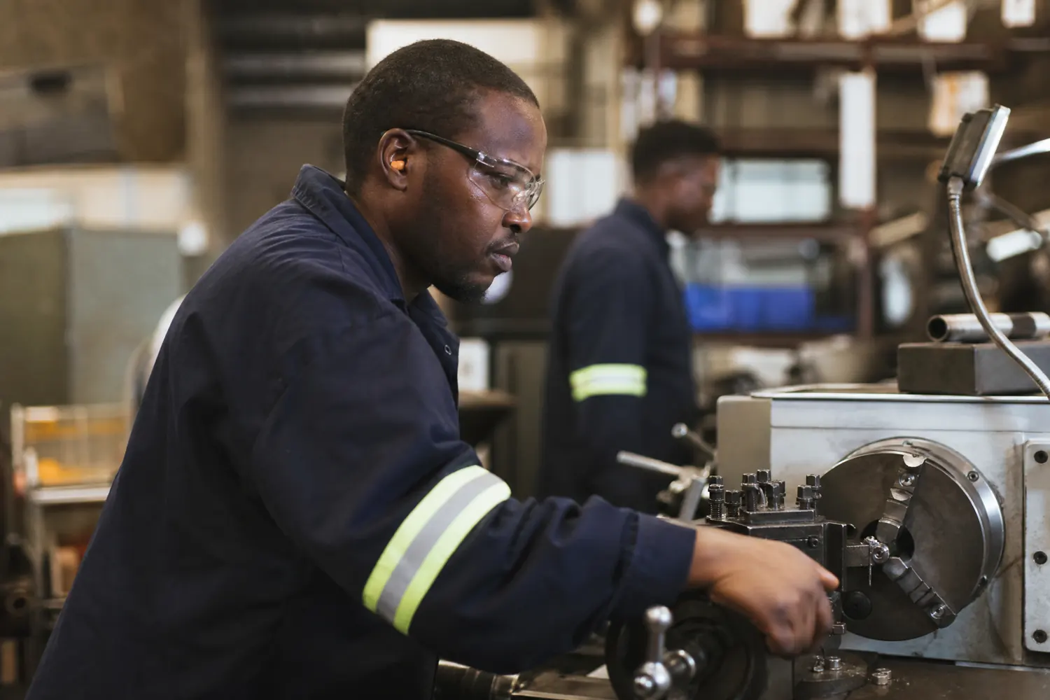 An operator in PPE and safety glasses works at a machine on a factory floor, a colleague at another station behind him.