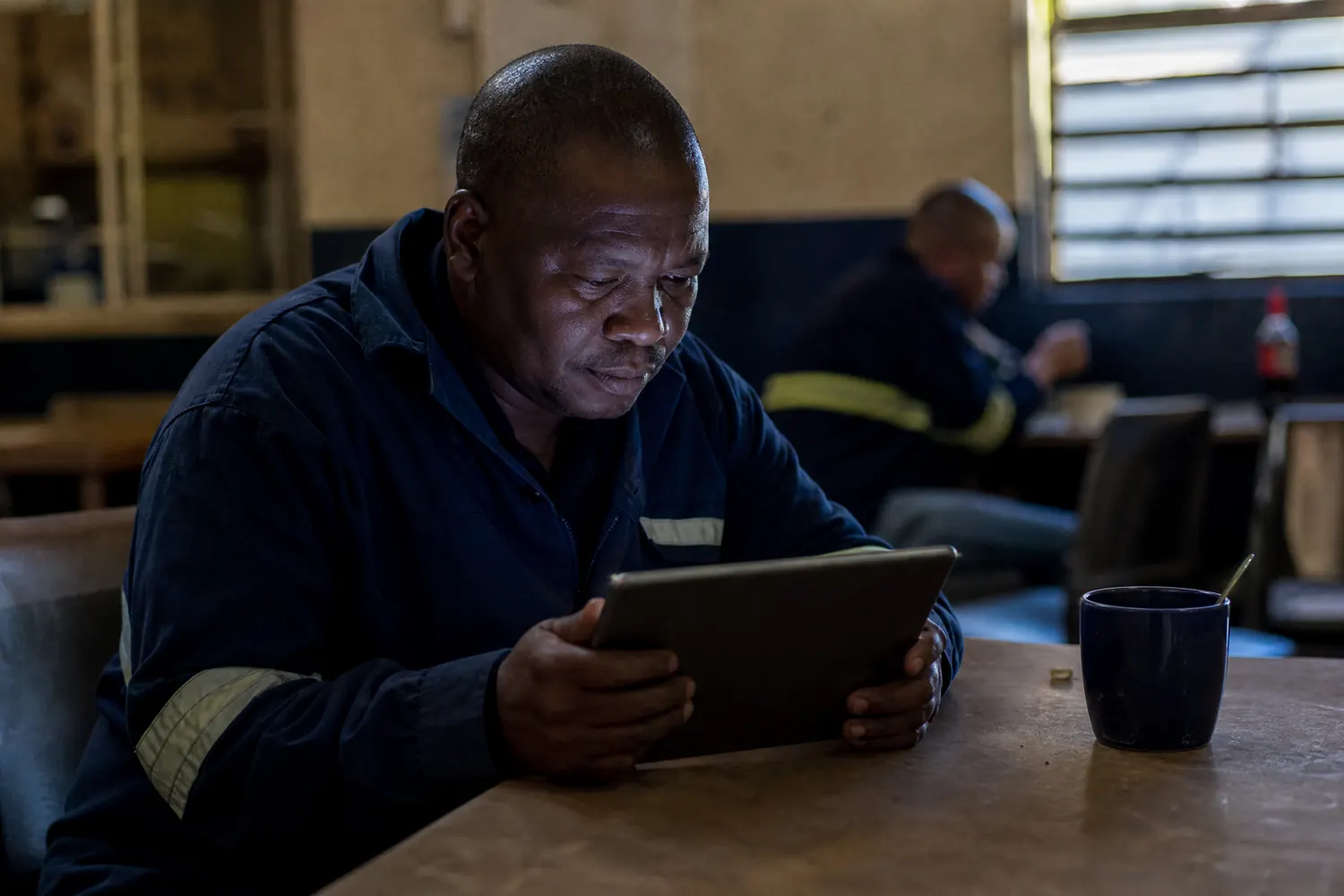 A driver in a depot break room studies an online course on a tablet, screen light on his face.