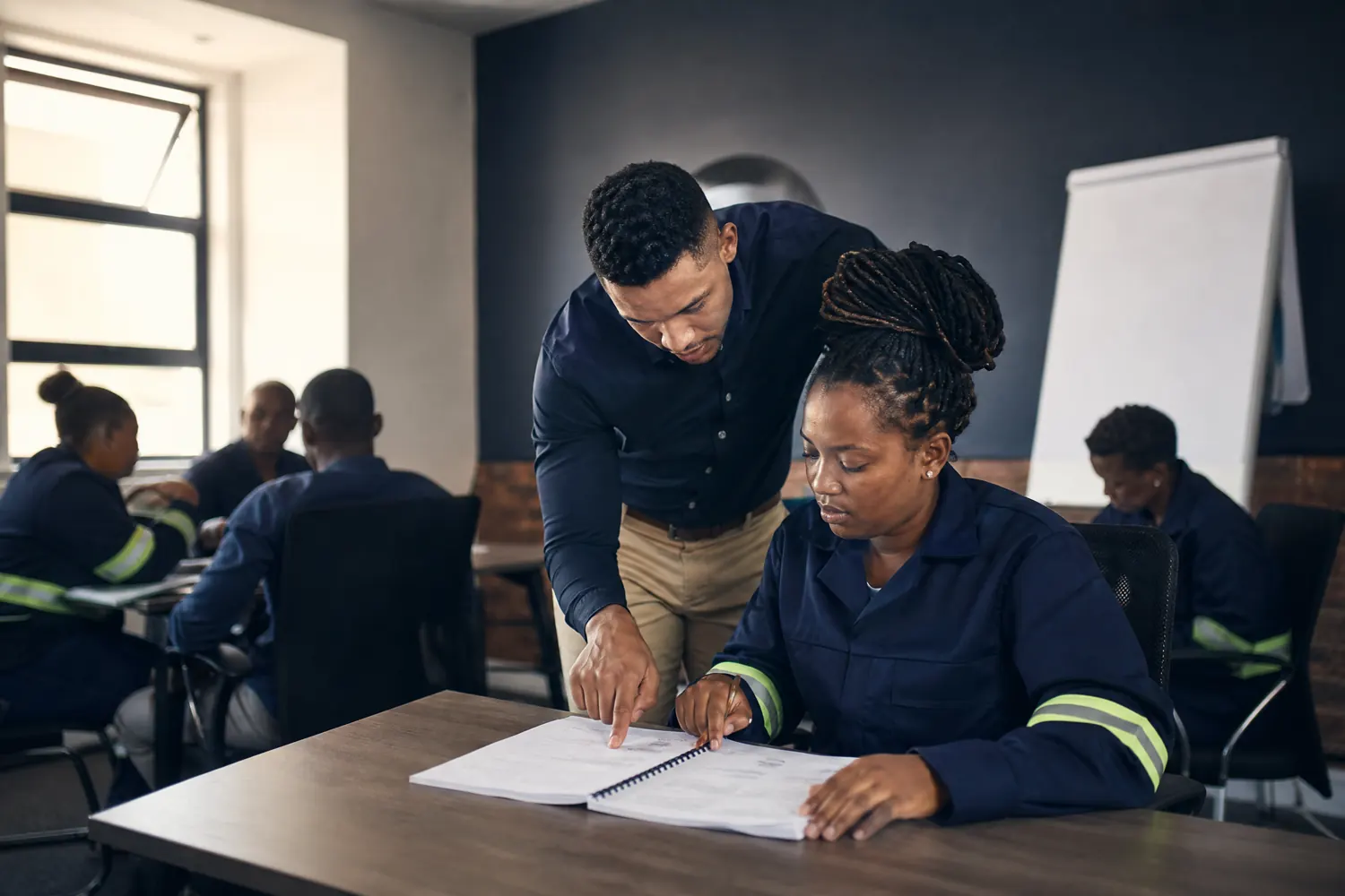 A facilitator points out a detail in a workbook to a seated learner during a training session, other learners at desks behind them.